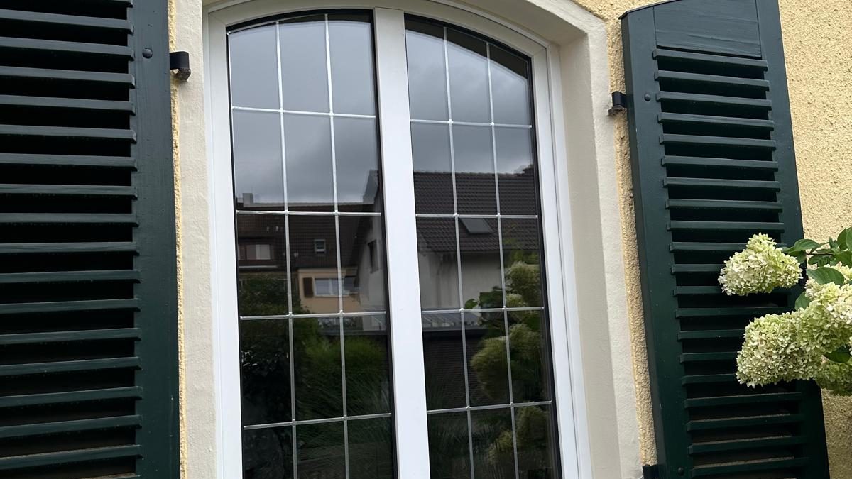 Weißes Rundbogenfenster mit Sprossen zwischen dunkelgrünen Klappläden